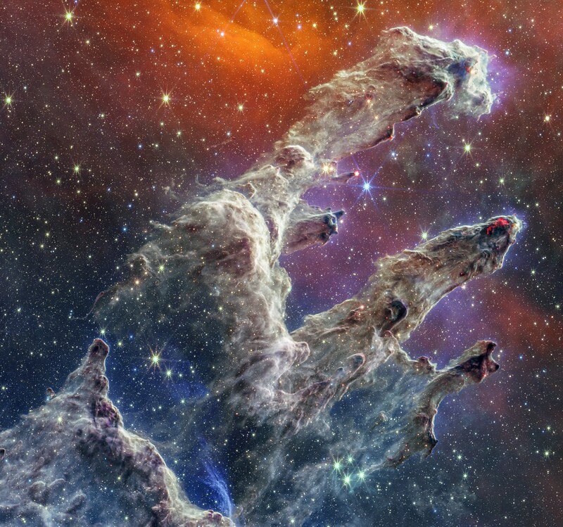 Picture from James Webb telescope: "pillars of creation". At the bottom left of this almost-square image are the thickest regions of brown and purple gas and dust. There are many layers of semi-transparent gas and dust overlaying one another. A peak rises about a third of the way from the bottom, and becomes far darker with two bright red areas toward the tip, which look like lava. The dust becomes sheer about halfway up the image. There’s a slight gap in the dust, which allows the multi-colored background to come into view. The background is blue at bottom left, orange at top center, and hazy pink in regions right outside the pillars. The light brown pillars continue, taking the shape of a shoulder at the base, with three prominent columns rising out toward the upper right. The top left pillar is the largest and widest. The peaks of the second and third pillars are set off in darker shades of brown and have transparent purple haze just outside them.